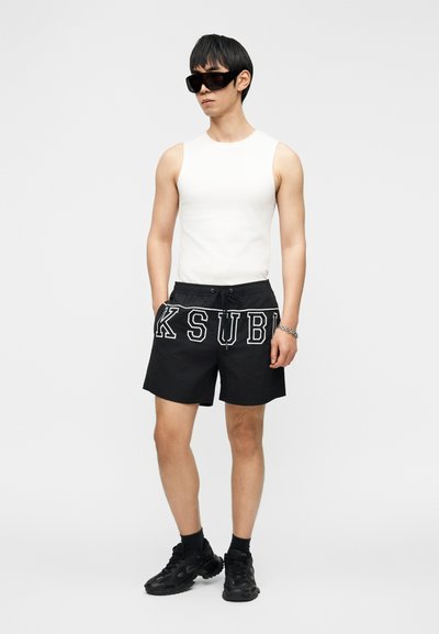 Vyras, dėvintis juodus didelius akinius nuo saulės, baltą berankovį viršutinį drabužį, juodus šortus su didelėmis baltomis raidėmis, juodas kojines ir juodus sportinius batelius, stovintis.