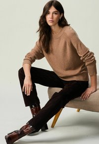 Tan pletený svetr s žebrovanými manžetami přes černé manžestrové kalhoty. Modelka má na sobě hnědé a černé lodičky a sedí na béžové židli.
