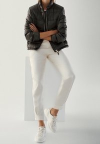 Personne portant une doudoune noire, un pantalon blanc et des baskets blanches, debout les bras croisés devant un fond gris clair.
