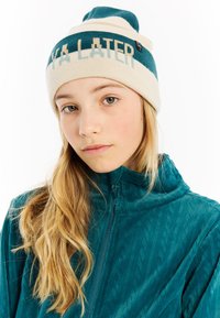 Jong persoon met lang blond haar, gekleed in een teal ritsvest en een beige en teal beanie met de tekst "LATER".