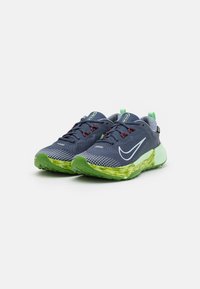 Baskets de sport navy avec une tige en mesh, semelle texturée verte et jaune, accents rouges sur les lacets, et logo Nike en swoosh réfléchissant.