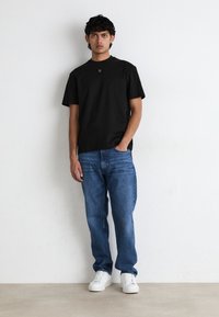 Svart t-skjorte med korte ermer og rund hals, kombinert med blå denimjeans og hvite joggesko. Modellen står mot en nøytral bakgrunn.
