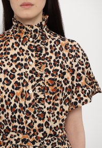 Beige leopardmønstret skjorte med korte ærmer, yndig krave og hvide knapper, med varierende mønstre i sorte og orange nuancer.