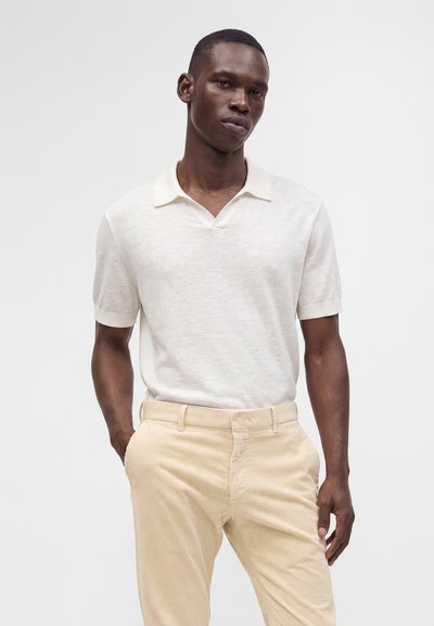 Homme portant un polo blanc à manches courtes rentré dans un pantalon en velours côtelé beige clair, les mains dans les poches, sur un fond uni.