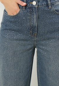 Jeans en denim à jambe large, de couleur bleu foncé et ornés de clous métalliques dispersés créant un motif texturé. Design classique à cinq poches.