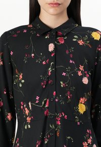 Svart kjole med blomsterbroderi i rosa, gult og grønt. Har krage, knappelukking og lange ermer. Myk stofftekstur.