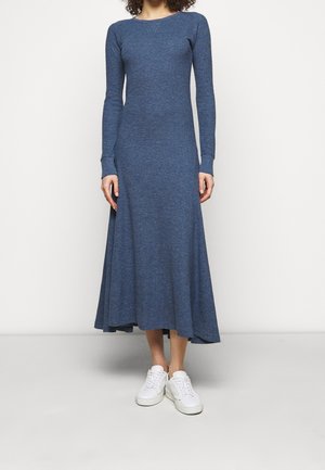 Person trägt ein langärmeliges, blaues Strick-Midikleid kombiniert mit weißen Sneakern und steht vor einem einfarbigen weißen Hintergrund.