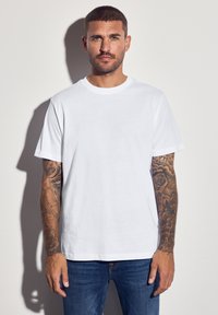 Bílý bavlněný tričko s kulatým výstřihem a krátkými rukávy, v kombinaci s modrými džíny. Má viditelné tetování na rukou a neutrální pozadí.