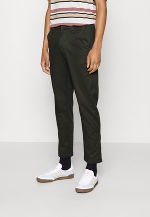 Green cotton chinos with a straight fit and side pockets. Paired with white sneakers featuring beige accents and a striped short-sleeve shirt.
