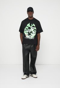 Tricou negru cu grafică de cranii și flăcări în verde, asociat cu pantaloni negri din piele ecologică și adidași albi cu dungi închise.