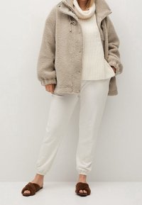 Beige fluffy jakke med høj krave, hvid strikket sweater, hvide tapered bukser og brune fuzzy sliders. Glatte teksturer overalt.