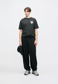 Černé bavlněné tričko s grafikou na levé straně hrudi, doplněné volnými černými kalhotami a šedými teniskami, drží černou čepici.