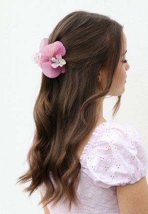 Donna con lunghi capelli castani ondulati, acconciati con una grande molletta a forma di orchidea rosa, che indossa una maglia rosa chiaro con maniche a sbuffo traforate, rivolta verso destra su sfondo bianco.