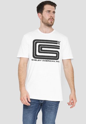 Weißes T-Shirt mit kurzen Ärmeln, das ein schwarzes Grafikdesign mit dem Buchstaben "S" und "SHELBY AMERICAN, INC." darunter zeigt. Lässiger Schnitt.