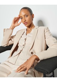 Beige linen suit with a fitted blazer and matching top, featuring rounded buttons and a relaxed fit. Model seated, hand resting on chin.