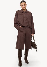 Chemise surdimensionnée en daim marron avec col, associée à un pantalon culotte assorti. Modèle portant des bottes marron brillantes montantes jusqu'aux genoux et tenant un sac à main.