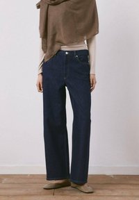 Persona que lleva jeans rectos azul oscuro, camiseta beige de manga larga, chal marrón y zapatos planos beige, de pie sobre un suelo de madera frente a una pared lisa.