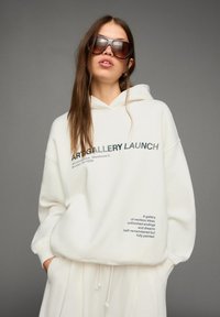 Jonge vrouw in een oversized witte hoodie met de tekst "ART GALLERY LAUNCH", bijpassende witte broek en grote bruine zonnebril tegen een grijze achtergrond.