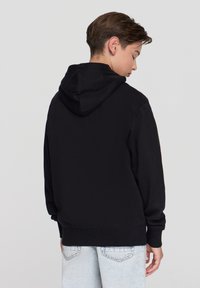 Felpa nera con cappuccio, maniche lunghe, polsini e vita a costine. Tessuto liscio, design semplice, indossata con denim chiaro.