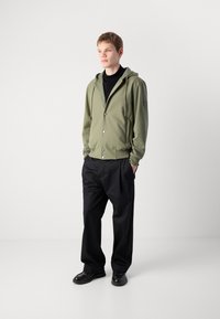 Joven de pie con las manos en los bolsillos, vestido con una chaqueta con capucha verde oliva, un cuello de tortuga negro, pantalones anchos negros y zapatos negros, contra un fondo blanco.