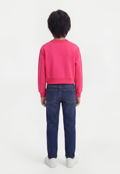 Παιδί που στέκεται με την πλάτη, φορώντας μια φωτεινή ροζ φούτερ, σκούρα μπλε τζιν και λευκά αθλητικά παπούτσια σε λευκό φόντο.