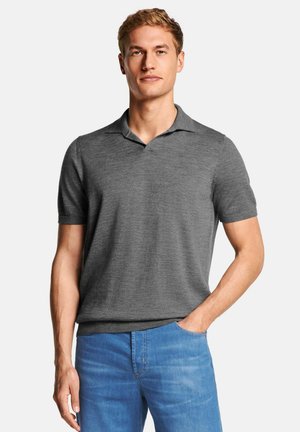 Uomo che indossa una polo grigia a maniche corte e jeans blu, in piedi con una mano in tasca contro uno sfondo semplice.