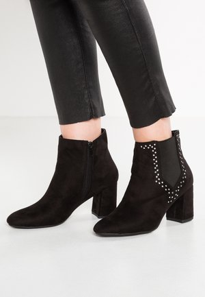 Zwarte enkellaarsjes van suède met blokhakken, zijrits en decoratieve zilveren studs, gedragen bij korte zwarte broek.