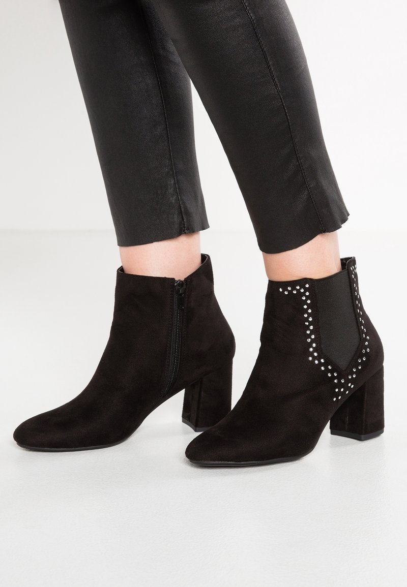Bottines en daim noir à talons carrés, fermeture éclair latérale et clous décoratifs en argent, portées avec un pantalon noir court.