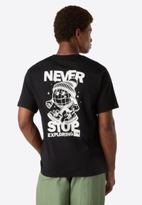 Man wearing black t-shirt with white graphic of walking globe in winter hat, text "Never Stop Exploring," and The North Face logo on back.