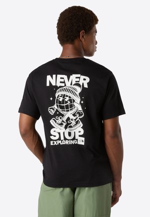 Mand iført sort T-shirt med hvid grafik af en gående globus i vinterhat, tekst "Never Stop Exploring," og The North Face-logo på ryggen.