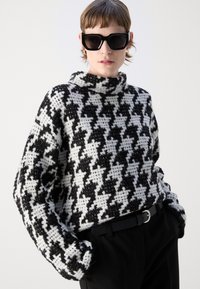Black and white houndstooth-patterned knitted sweater with a high neck and loose fit, paired with a black belt and black pants.