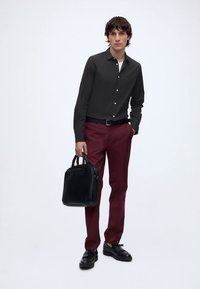 Young man wearing a black buttoned shirt, burgundy pants, black belt, and black shoes holding a black leather briefcase against a plain background.