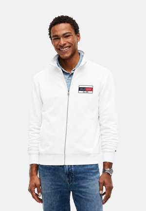 Hombre sonriente con chaqueta blanca con cremallera y logo de Hilfiger, camisa azul clara, vaqueros azules y reloj de pulsera plateado en la muñeca izquierda.
