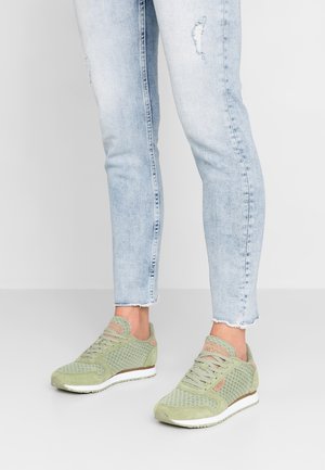 Personne portant un jean skinny bleu clair déchiré et des baskets en suède vert et mesh, debout sur une surface blanche.