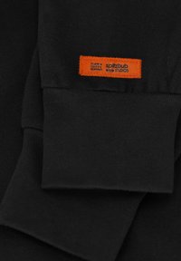 Zwarte sweatshirtstof met geribbelde manchetten. Bevat een oranje label dat op de manchette is gestikt met coördinaten en de merknaam "spitzbub STUDIOS."