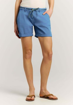Vrouw draagt lichtblauwe opgerolde shorts met een patroonkoord, beige tanktop en bruine slippers, handen ontspannen langs het lichaam.