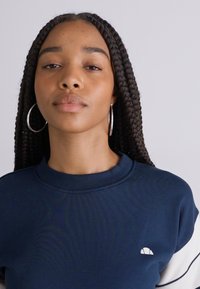 Sweatshirt bleu marine avec un col rond, un col côtelé et des accents blancs sur les épaules. Présente un petit logo sur la poitrine.