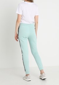 Mátově zelené tepláky s černými bočními pruhy, zúžený střih, materiál s podílem bavlny, v kombinaci s jednoduchým bílým tričkem s krátkým rukávem.