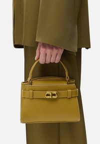 Kleine gele handtas van textuurleer, met een dubbele gespsluiting en een korte bovenhandgreep. Draagt een groene outfit.