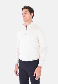 Maglione beige chiaro a zip corta con maniche lunghe, polsini e orlo a coste, e una zip di colore contrastante; abbinato a pantaloni scuri.