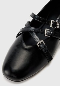 Black synthetic leather shoe with a rounded toe, featuring two crossing straps with silver buckles across the midfoot. Smooth texture.
