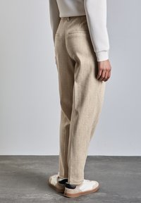 Beige, strukturierte, hochgeschnittene Hose mit zwei Gesäßtaschen, kombiniert mit weißen und grauen Sneakers, aus einer Rückansicht gezeigt.