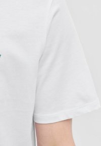 Hvid bomuld t-shirt med korte ærmer, der har et enkelt design. Glat tekstur med en simpel kant og et nærbillede af ærmet og armen.