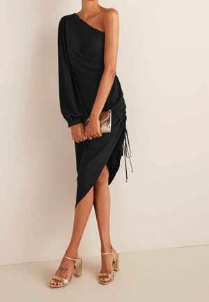 Frau trägt ein schwarzes Ein-Schulter-Kleid mit geraffter Seite, hält eine bronzefarbene Clutch, kombiniert mit goldenen Sandalen mit klobigem Absatz und Knöchelriemen.