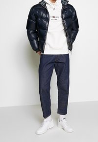 Giubbotto imbottito navale con texture a nido d'ape, felpa bianca con logo, jeans scuri e sneakers bianche. Include chiusura con zip e colletto a fantasia.