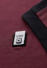 Bordeaux tričko s černým výstřihem, s textilním štítkem se značkou "ombre menswear" a akcentem ve čtvercovém designu.