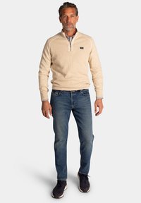 Beige pullover met kwartrits, gestructureerde stof, marineblauwe jeans, blauwe sneakers. De pullover heeft een klein logo op de borst en geribbelde manchetten.