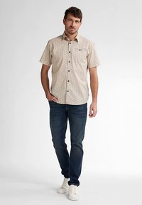 Beige lyhythihainen paita, jossa tummat pilkut, napillinen etuosa ja rintatasku, yhdistetty tummansinisiin farkkuihin ja valkoisiin lenkkareihin.