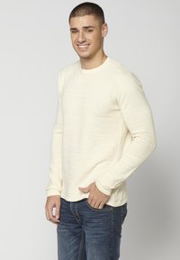 Cremefarbenes Langarmshirt mit geripptem Muster, rundem Ausschnitt und enganliegenden Ärmeln, kombiniert mit blauen Jeans. Einfaches und modernes Design.