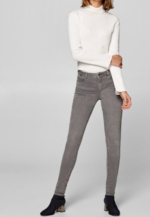 Femme portant un pull col roulé blanc à manches longues, un jean skinny gris et des bottines noires, debout devant un fond gris uni.